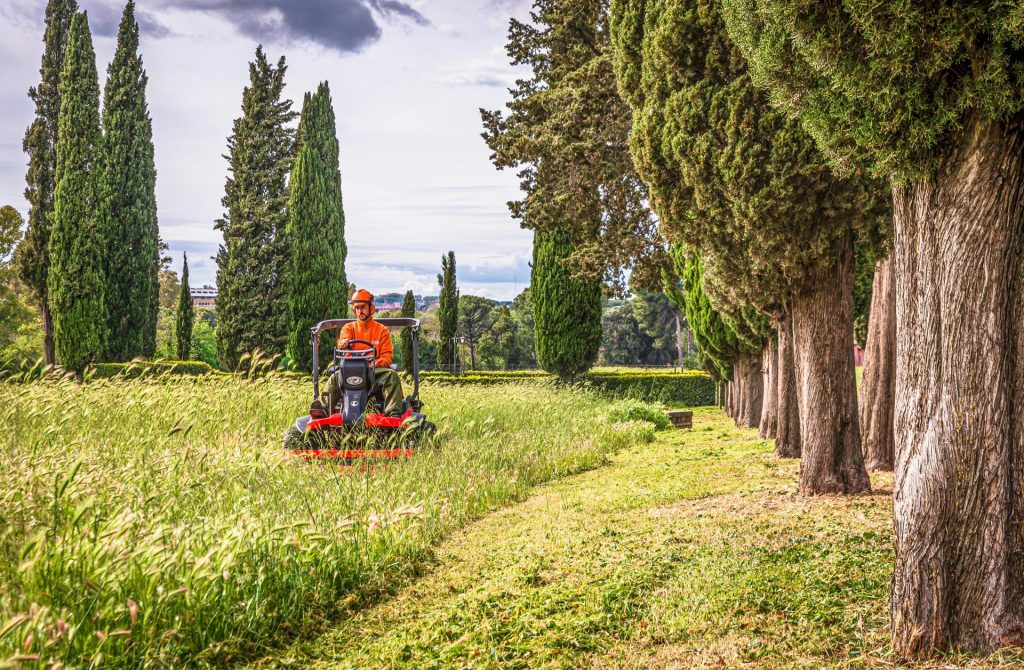 Ein Mann bedient einen Rasenmäher in einer malerischen italienischen Landschaft mit Zypressen.